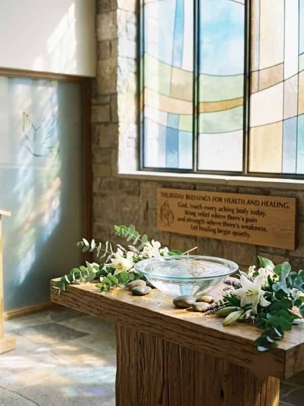 An image featuring a clear glass bowl filled with water resting on a rustic wooden table in a serene chapel setting, surrounded by white lilies and green foliage. A wooden plaque on the stone wall behind it bears the text, "THURSDAY BLESSINGS FOR HEALTH AND HEALING. God, touch every aching body today. Bring relief where there's pain and strength where there's weakness. Let healing begin quietly." Soft sunlight filters through stained-glass windows onto the scene.