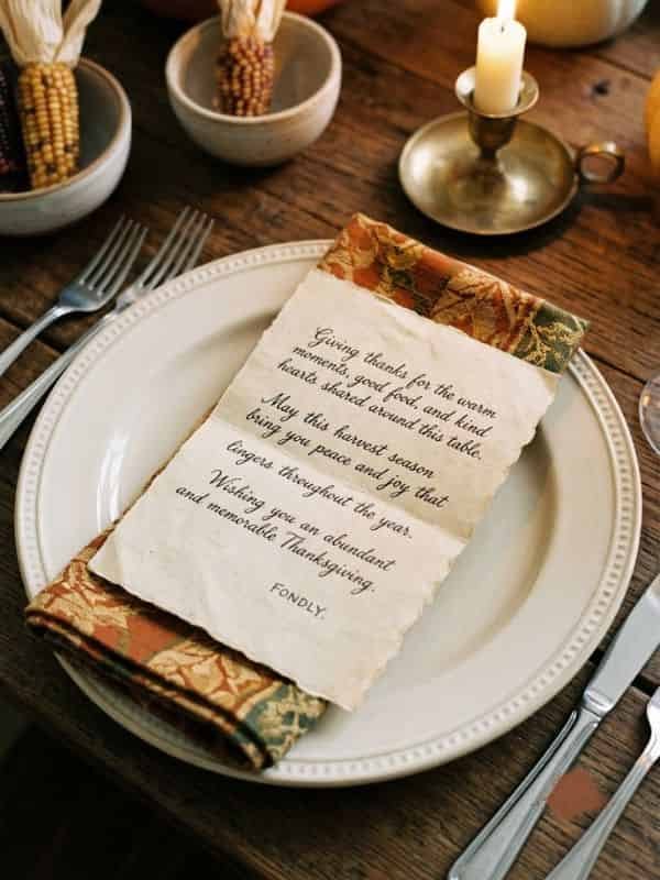 A rustic Thanksgiving table setting featuring a handwritten note placed on a folded patterned napkin atop a ceramic dinner plate, alongside a lit brass candlestick and wooden bowls filled with decorative corn.